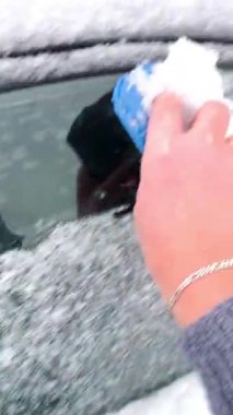 Mavi kazayağı olan gri kazaklı bir kadın, bir kış sabahı siyah bir arabanın camından kar ve don tabakasını çıkarır..