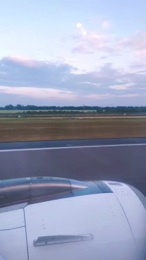 Uçak penceresinden podyumdan aşağı inerken ve havalanırken bir görüntü. Çerçevede uçak motoru, yeşil bir tarla ve şafak pembesiyle bezenmiş bir gökyüzü var..