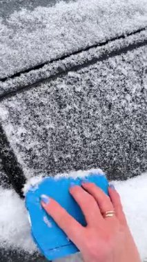 Mavi kazayağı olan gri kazaklı bir kadın, bir kış sabahı siyah bir arabanın camından kar ve don tabakasını çıkarır..