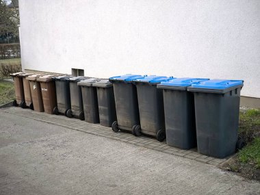 Binaların beyaz duvarı boyunca kahverengi, siyah ve mavi renkli çöp tenekeleri dizilmiştir. Ekoloji, atık geri dönüşümü ve kentsel yönetim