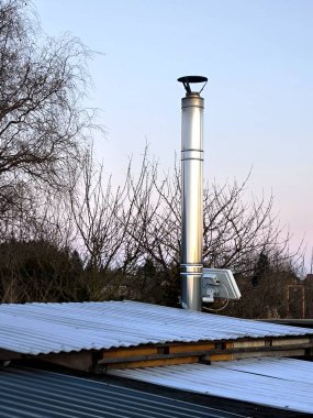 Oluklu bir çatıya paslanmaz çelik bir dikey boru monte edilir. Bağlayıcılar ve saptırıcı kapağı görünür durumda. Arkaplanda ağaç dalları ve açık bir gökyüzü var..