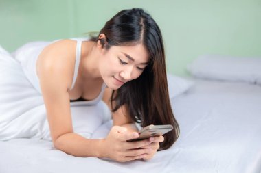 Güzel Asyalı kadın yatak odasında cep telefonu kullanıyor ve gülümsüyor..