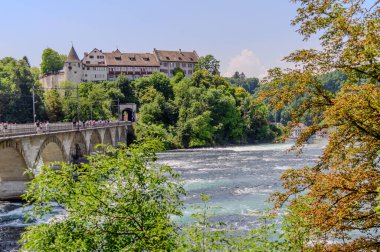 Arka planda Schloss Laufen kalesinin yer aldığı Ren Şelalesi 'nin (Rheinfall) panoramik manzarası. Yayalarla dolu taş bir köprü nehri boydan boya geçiyor ve yemyeşil alan manzarayı süslüyor..