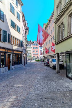 İsviçre, Zrich 'te canlı bir sokak sahnesi. Cobblestone caddesi kepenkli pencereleri ve dekoratif cepheleri olan tarihi binalarla dolu. Bir İsviçre bayrağı belirgin bir şekilde dalgalanıyor. Caddenin kenarına beyaz bir minibüs park etmiş..