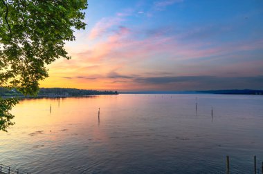 Constance Gölü (Bodensee) üzerinde sakin bir gündoğumu. Durgun su, pembe ve turuncudan maviye dönüşen gökyüzünün canlı renklerini yansıtır. Sudan birkaç tahta direk yükselir ve sahneye ince bir doku ekler..