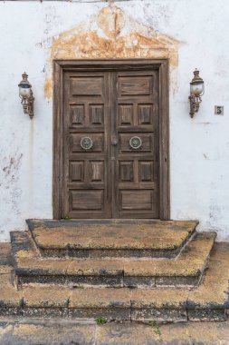 Rustic wooden door with ornate details, set into a weathered white wall with stone steps. Two lanterns flank the door, adding a touch of old-world charm. The scene evokes a sense of history and tranquility.