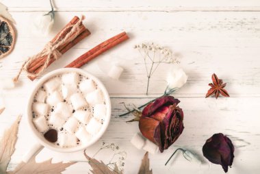 Bir fincan kremalı sıcak çikolata, tarçın çubukları, beyaz ahşap arka planda kurumuş gül.