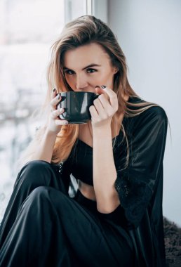 Biri siyah elbiseli beyaz bir kadın sabahları pencerenin yanındaki bardaktan su içer.