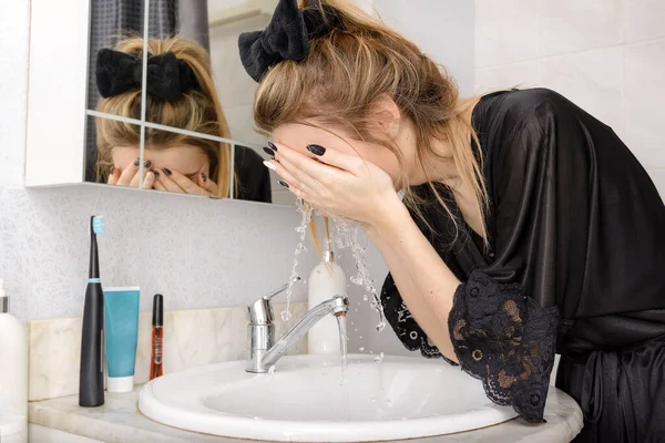 blonde woman in a black negligee washes her face in the bathroom