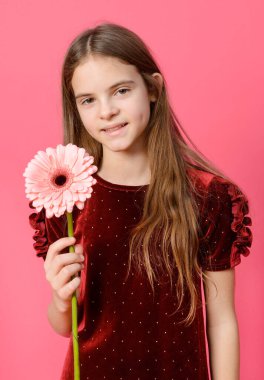 1 beyaz kız. 10 yaşında, kırmızı elbiseli. Elinde pembe bir Gerbera çiçeği var.