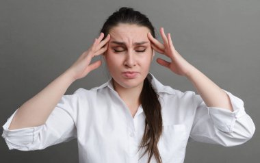 Beyaz gömlekli bir beyaz kadın ellerini gri arka planda tutuyor, başı ağrıyor.,