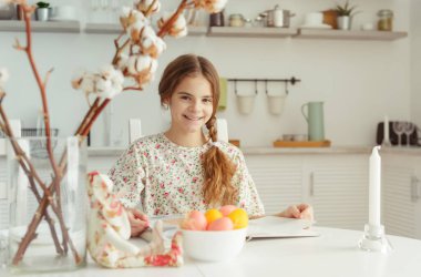  Avrupalı kız. 10 yaşında. Mutfakta, masada. Elinde sarı, pembe Paskalya yumurtaları var.