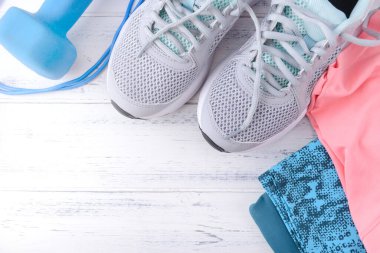 Gri spor ayakkabılar, mavi halterler, pembe spor giysiler, beyaz ahşap arka planda.