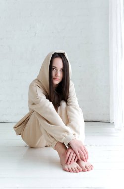 brunette European girl in sweatpants and a hoodie sits on a white wooden floor in a bright room clipart