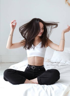 Beyaz bluzlu esmer kız. Parlak bir odada uzun saçlı yatakta oturuyor.