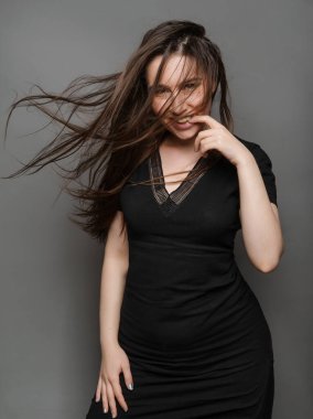 Gri arka planda gülümseyen siyah elbiseli, uzun saçlı güzel ve çekici genç bir kadın.
