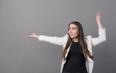 Beyaz ceketli, uzun saçlı genç bir kadın ellerini sallıyor ve gri arkaplandaki duygulara şaşırıyor.