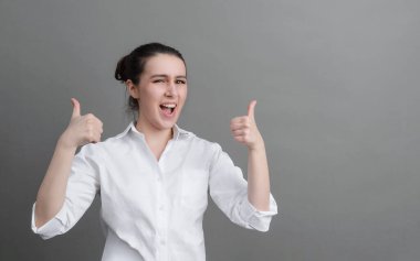 Beyaz gömlekli beyaz kadın mutlu ve gri arka planda bir sınıf gösteriyor.