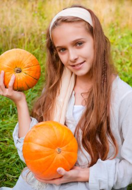 1 beyaz kız 11 yaşında, elinde yeşil arka planda 2 tane turuncu balkabağı tutuyor.