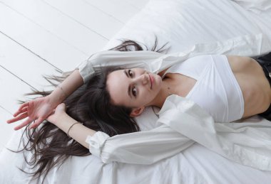 Beyaz tişörtlü, uzun saçlı, beyaz, güzel esmer kadın. Yatakta uzanıyor ve gülüyor.