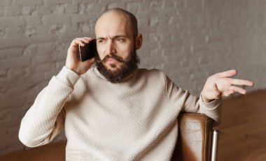 Beyaz, kel, sakallı adam odada telefonla konuşuyor.