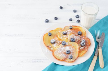 Tabakta yaban mersinli krep ve pudra şekeri, masada bir bardak süt, bıçak, mavi peçete, çatal, üst manzara.