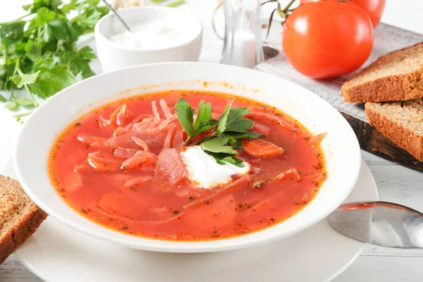 Ekşi kremalı kırmızı borş ve beyaz bir tabakta otlar, ekmek, beyaz tahta bir masada kaşık