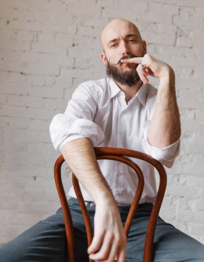 Beyaz, kel, sakallı ve bıyıklı bir adam. Odada bir sandalyede oturuyor.