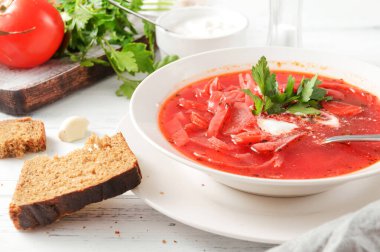 Kırmızı çorba, ekşi kremalı borş ve beyaz tabakta maydanoz, bir kaşık, beyaz ahşap bir masada ekmek.