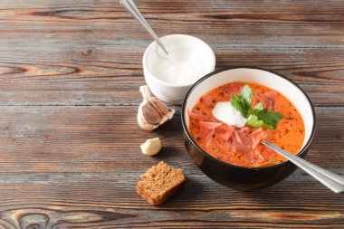 Ekşi kremalı ve maydanozlu kırmızı borş, ekmek, kaşık, sarımsak, koyu ahşap bir arka planda.