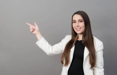 Uzun saçlı, beyaz ceketli genç bir kadın gülümsüyor, gri bir arka planı işaret ediyor.