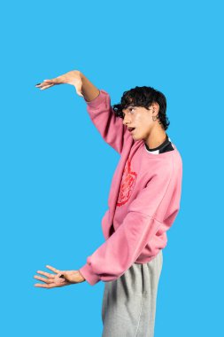 Genç adam mavi bir arka planda pembe bir kazakla şakacı poz veriyor.