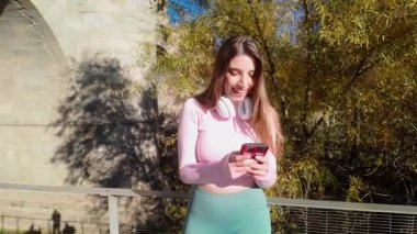Güneşli bir günde, köprüde cep telefonunu kullanan gülümseyen atletik bir kadın.