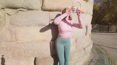 Formda bir kadın atletizm kıyafetleri içinde kulaklıklarla müziğin keyfini çıkarıyor ve dışarıda taş bir duvara karşı dans ediyor.