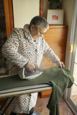 Üst düzey kadın ev işi yapıyor, evde buharlı ütüyle yeşil pantolonları ütülüyor.