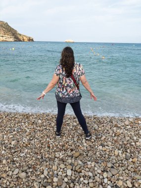 Çakıl taşı plajında duran bir kadın, sakin okyanusa ve uzak ufka bakıyor.