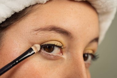 Makyaj fırçasıyla göz kapağına sarı göz farı süren bir kadın, güzellik rutinine odaklanıyor.