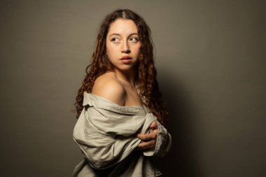 Kıvırcık saçlı, omuzları açık bir gömlek giyen genç bir kadın stüdyo ortamında gözlerini kaçırıyor.