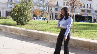 Şık profesyonel siyahi kadın şehirde yürürken akıllı telefonuyla konuşuyor.