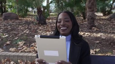 Şehir parkında bir bankta otururken video araması için tablet kullanan neşeli genç siyahi kadın.