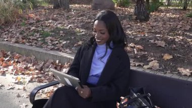 Sonbaharda bir bankta otururken dijital tablet üzerinde çalışan kendine güvenen siyahi iş kadını