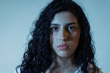 Kıvırcık saçlı, yüzünde ciddi bir ifade olan genç bir kadın kameraya bakıyor.