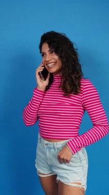 Neşeli kıvırcık saçlı kadın mavi bir arka planda akıllı telefonuyla sohbet ediyor ve gülümsüyor.