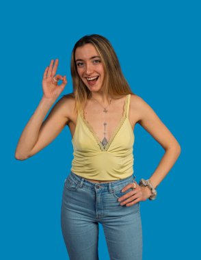 Gülümseyen genç bir kadın, mavi bir arka planda duruyor, iyi bir el hareketi gösteriyor.