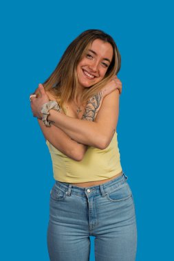 Kendini kucaklarken gülümseyen genç bir kadın, mavi bir arka planda kendine olan güvenini ve sevgisini ifade ediyor.