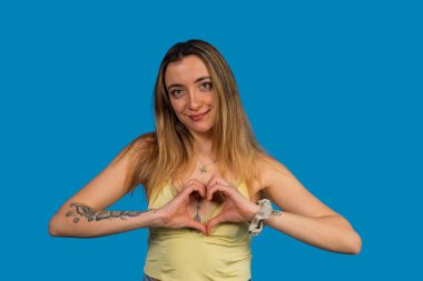 Genç bir kadın gülümsüyor ve elleri mavi bir arka planda kalp şekli oluşturuyor.