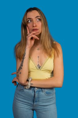 Dövmeleri olan genç bir kadın dikkatlice düşünüyor, mavi bir arka plana bakıyor.