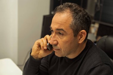 Siyah kazaklı bir adam profesyonel bir ortamda telefonla konuşuyor.