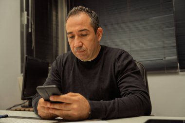 Orta yaşlı bir adam masada oturuyor, dikkatle akıllı telefon ekranını kullanıyor.