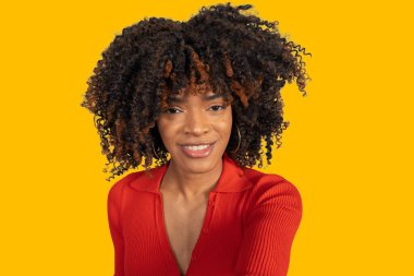 Genç siyah kadın gülümsüyor, kameraya bakıyor, sarı arka planda duruyor.